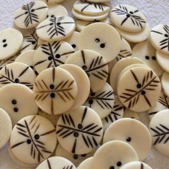 A pile of off white bone buttons that have 2 holes. The buttons are carved and the carving has a dark brown paint applied.