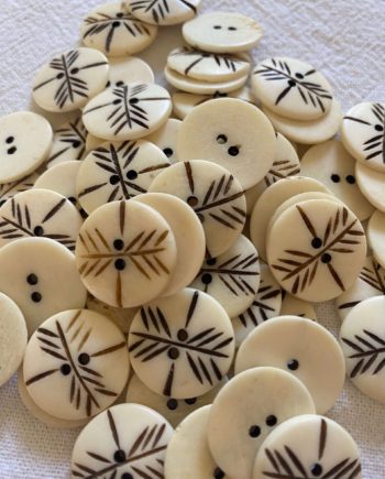 A pile of off white bone buttons that have 2 holes. The buttons are carved and the carving has a dark brown paint applied.