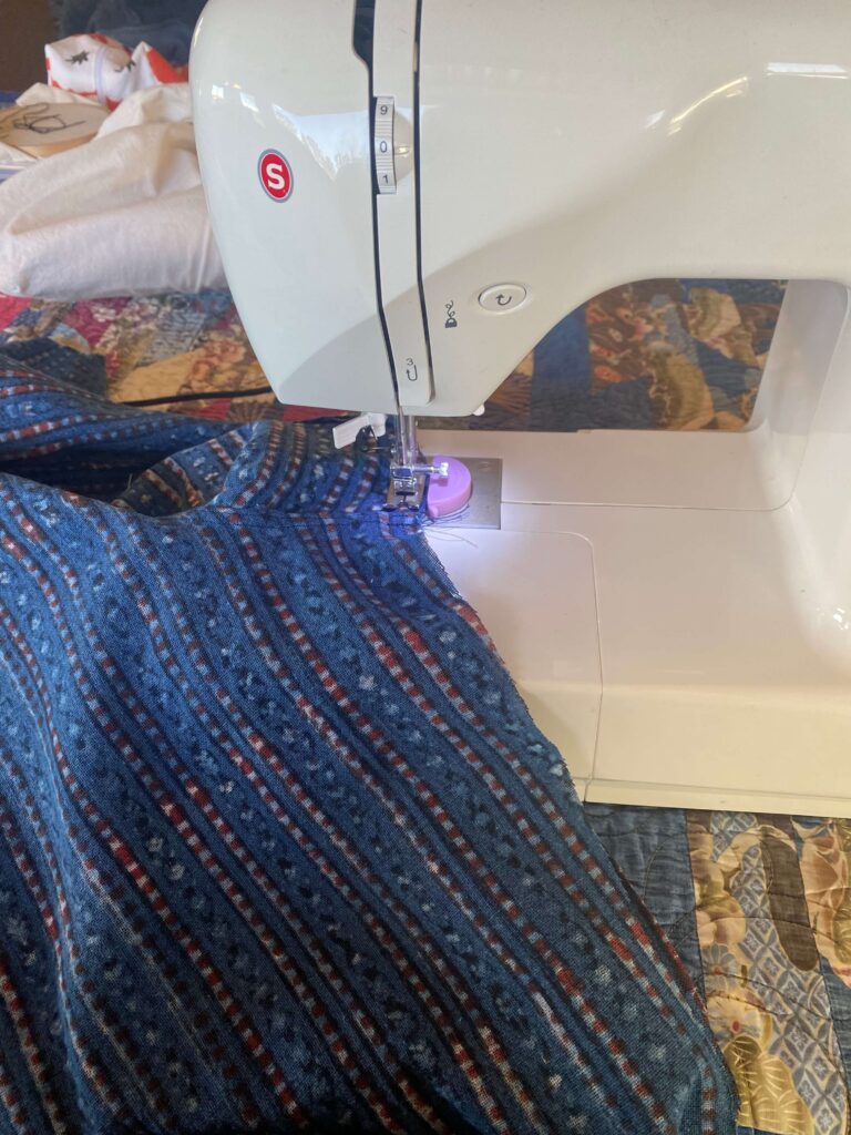 Person sits at a sewing machine, guiding blue patterned fabric under the needle as it sews. A purple bobbin area is visible under the machine.