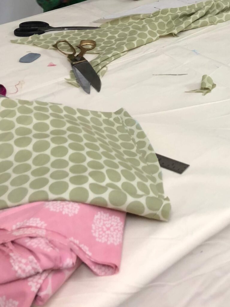 Scissors resting on a sewing table beside green patterned fabric scraps and pink fabric pieces.
